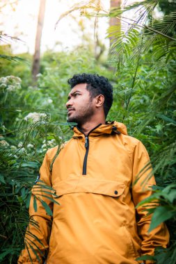 Sarı ceket giyen bir adam yoğun bitki örtüsü ve tropikal bitkilerle çevrili yemyeşil bir ormanı keşfeder. Güneş ışığı ağaçların arasından süzülüp sakin ve maceracı bir atmosfer yaratır..