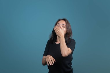 Endonezyalı bir kadın tek eliyle ağzını kapatarak neşeyle güler. Siyah giyinmiş ve mavi bir arka plana yaslanmış, doğal gülüşü gerçek mutluluğu yakalıyor..