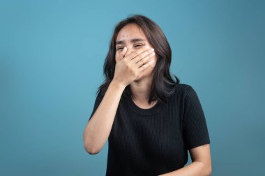 Endonezyalı bir kadın tek eliyle ağzını kapatarak neşeyle güler. Siyah giyinmiş ve mavi bir arka plana yaslanmış, doğal gülüşü gerçek mutluluğu yakalıyor..