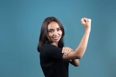 Endonezyalı bir kadın güçlü bir poz verirken kendine güvenerek gülümsüyor. Siyah bir gömlek giyiyor ve mavi bir arka plana karşı duruyor. Gücü, gücü ve pozitifliği simgeliyor..