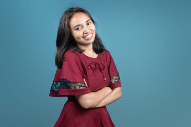 Kırmızı elbiseli, kolları çapraz gülümseyen Endonezyalı kadın, mavi arka planda güvenle poz veriyor. Çeşitlilik, yaşam tarzı, portre ve moda içeriği için ideal.