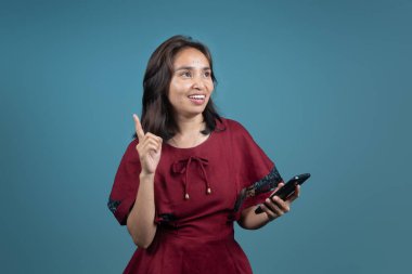 Kırmızı elbiseli mutlu Endonezyalı kadın akıllı telefon tutuyor ve gülümseyerek işaret ediyor. Teknoloji, yaşam tarzı, iletişim ya da fikir konsepti görselleri için harika.