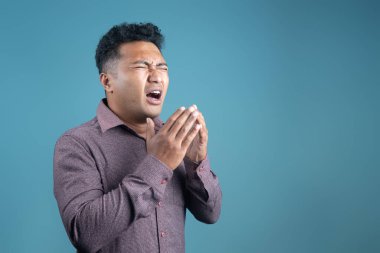 Kestane rengi gömlekli genç adam gözlerini kapatıyor ve yüzünü tutarak hapşırıyor gibi görünüyor. Mavi arka planda stüdyo çekimi. Soğuk algınlığı, grip, alerji veya hastalık reaksiyonu konsepti.