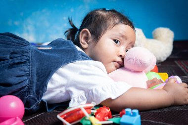 Asyalı küçük bir kız, oyuncaklarla çevrili bir oyuncak ayıya sarılarak yüzüstü yatıyor. Yumuşak bir gülümsemesi var. Yorgunluk belirtileri var. Oyun zamanından sonra sessiz bir anı yakalıyor..