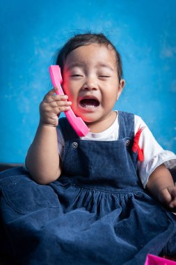 Endonezyalı bir çocuk pembe bir oyuncak telefonu tutarken kafası karışmış görünüyor, gerçek bir aramayı belirsiz bir sesle taklit ediyor. İfade dolu yüzü her şeyi anlatıyor. Ne dedin??