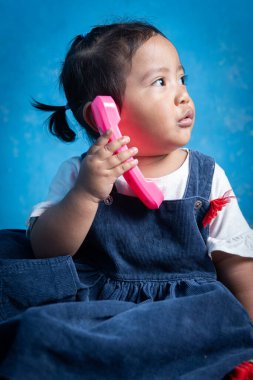 Sevimli çocuk kulağına pembe bir oyuncak telefon tutarken yana doğru merakla bakıyor. Yumuşak mavi bir arka planı ve neşeli bir ifadesi olan bir stüdyoda yakalandı..