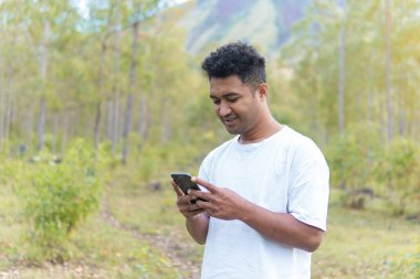 Beyaz gömlek giyen Asyalı bir Endonezyalı bir adam ormanın ortasında duruyor ve modern hayatı doğanın sükunetiyle harmanlayan bir telefon görüşmesi yapıyor..