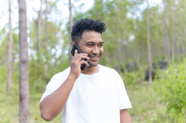 Beyaz gömlek giyen Asyalı bir Endonezyalı bir adam bir ormanın ortasında duruyor ve doğanın sükunetiyle modern hayatı karıştıran bir telefon görüşmesi yapıyor..