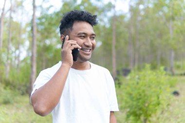 Beyaz gömlek giyen Asyalı bir Endonezyalı bir adam ormanın ortasında duruyor ve modern hayatı doğanın sükunetiyle harmanlayan bir telefon görüşmesi yapıyor..