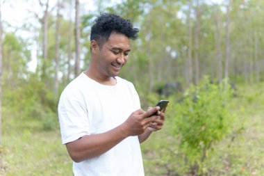 Beyaz gömlek giyen Asyalı bir Endonezyalı bir adam bir ormanın ortasında duruyor ve doğanın sükunetiyle modern hayatı karıştıran bir telefon görüşmesi yapıyor..