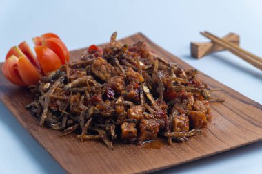 Geleneksel baharatlı Endonezya yemeği, dilimlenmiş domateslerle süslenmiş ahşap bir tabakta servis edilen kızarmış ançüez ve tempeh. Mutfak, yemek ve kültürel içerik için mükemmel..