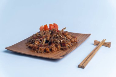 Geleneksel baharatlı Endonezya yemeği, dilimlenmiş domateslerle süslenmiş ahşap bir tabakta servis edilen kızarmış ançüez ve tempeh. Mutfak, yemek ve kültürel içerik için mükemmel..