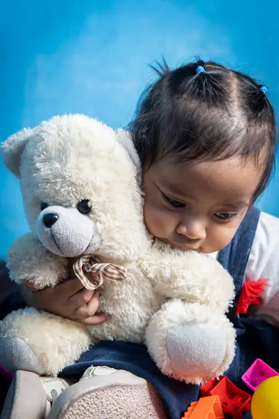 Sevimli bebek evde otururken yumuşak bir oyuncak ayıya sevgiyle sarılıyor. Mavi bir arka planda yakalanan bu yürek ısıtan an saf masumiyet ve şefkat göstergesidir..