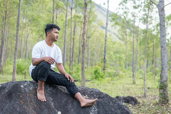 Asyalı bir Endonezyalı, bereketli ormanlarla çevrili büyük bir kayanın üzerinde sakince oturur. Huzur, yalnızlık ve doğayla olan bağını yakalar..