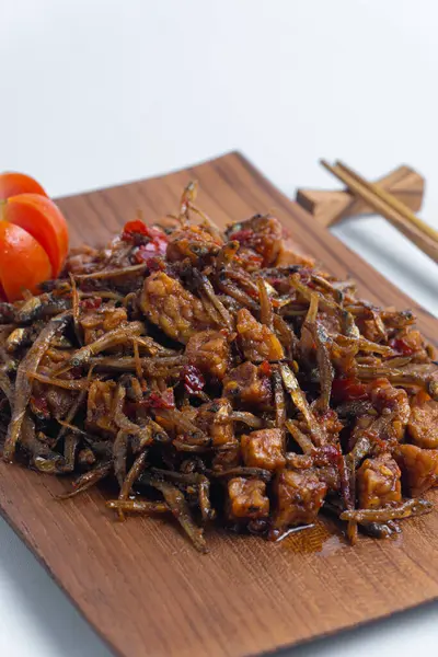Geleneksel baharatlı Endonezya yemeği, dilimlenmiş domateslerle süslenmiş ahşap bir tabakta servis edilen kızarmış ançüez ve tempeh. Mutfak, yemek ve kültürel içerik için mükemmel..