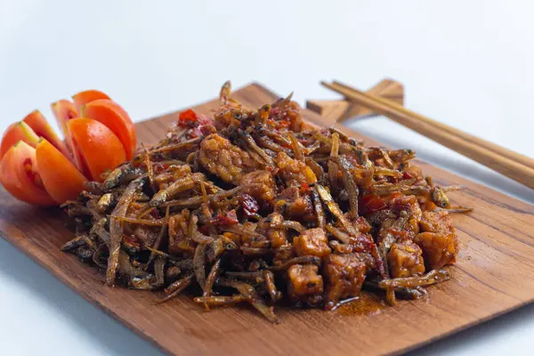 Geleneksel baharatlı Endonezya yemeği, dilimlenmiş domateslerle süslenmiş ahşap bir tabakta servis edilen kızarmış ançüez ve tempeh. Mutfak, yemek ve kültürel içerik için mükemmel..