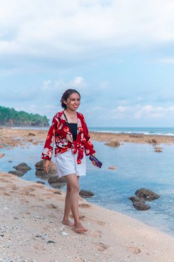 Güneşli bir tropikal plajda çıplak ayakla yürüyen mutlu bir kadın. Kırmızı çiçekli bir bluz ve beyaz şort giyiyor. Yeşil orman, kum ve kayaların arka planda olduğu doğal samimi bir an..