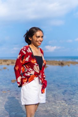Güneşli bir tropikal plajda çıplak ayakla yürüyen mutlu bir kadın. Kırmızı çiçekli bir bluz ve beyaz şort giyiyor. Yeşil orman, kum ve kayaların arka planda olduğu doğal samimi bir an..