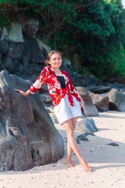 Genç Endonezyalı kadın tropik bir plajda büyük bir kayaya yaslanırken gülümsüyor. Kırmızı çiçekli bir gömlek ve beyaz bir şort giyerek doğayla çevrili güneşli günün tadını çıkarıyor..