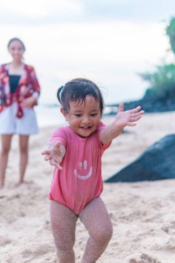 Pembe elbiseli neşeli bir çocuk, kolları kumla kaplı, tropik bir plajda neşeyle oynar. Arka planda bir kadın sıcak, samimi bir aile tatili havası katıyor..