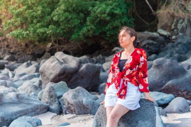 Endonezyalı bir kadın kumsaldaki bir kayanın üzerinde çıplak ayakla oturuyor, üzerinde çiçekli kırmızı bir bluz ve beyaz bir şort var. Doğal ortam, rahat duruş ve tropikal atmosfer boş zamanları ve tatil atmosferini ön plana çıkarıyor..