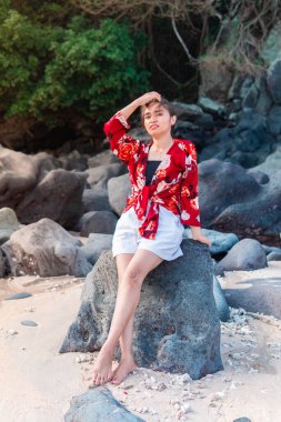 Endonezyalı bir kadın kumsaldaki bir kayanın üzerinde çıplak ayakla oturuyor, üzerinde çiçekli kırmızı bir bluz ve beyaz bir şort var. Doğal ortam, rahat duruş ve tropikal atmosfer boş zamanları ve tatil atmosferini ön plana çıkarıyor..