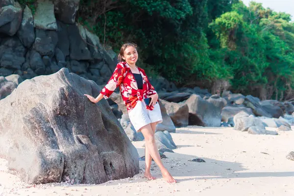 Genç Endonezyalı kadın tropik bir plajda büyük bir kayaya yaslanırken gülümsüyor. Kırmızı çiçekli bir gömlek ve beyaz bir şort giyerek doğayla çevrili güneşli günün tadını çıkarıyor..