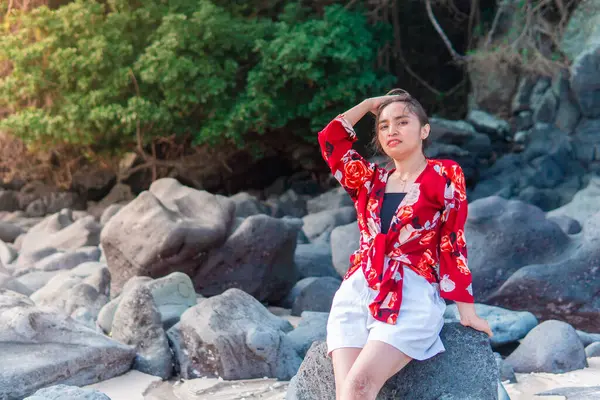 Endonezyalı bir kadın kumsaldaki bir kayanın üzerinde çıplak ayakla oturuyor, üzerinde çiçekli kırmızı bir bluz ve beyaz bir şort var. Doğal ortam, rahat duruş ve tropikal atmosfer boş zamanları ve tatil atmosferini ön plana çıkarıyor..