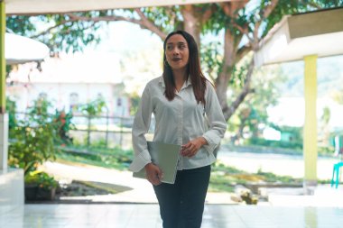 Ofis terasında elinde dizüstü bilgisayarla duran kendine güvenen Endonezyalı kadın. Arka plan yeşillik ve ağaçları gösteriyor, modern çalışmayla doğal ortam koşullarını birleştiriyor. Teknoloji ve yaşam tarzı temaları için harika.