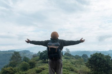Bereli ve sırt çantalı Endonezyalı bir adam bulutlu bir gökyüzünün altındaki yeşil bir yamaçta durarak kollarını ardına kadar açıyor. Özgürlüğün, minnettarlığın ve doğayla bağın güçlü bir ifadesi..