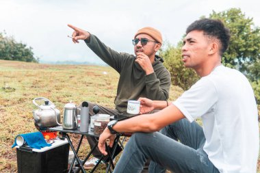 İki Endonezyalı adam kahve hazırlarken rahat bir ortamda birlikte gülüyorlar. Rahat bir kamp, kaynaşma ve çimenli bir arazide taşınabilir kahve takımlarıyla doğanın tadını çıkarma anı..