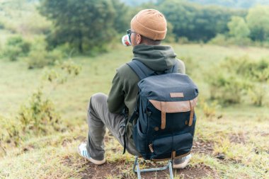Bir adam kamp sandalyesine oturmuş, sırt çantası ve bir fincan kahveyle huzurlu dağ manzarasına hayran. Bir anlık yalnızlık, yansıma ve doğa anlayışı..