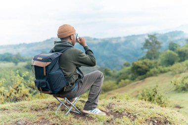 Bir adam kamp sandalyesine oturmuş, sırt çantası ve bir fincan kahveyle huzurlu dağ manzarasına hayran. Bir anlık yalnızlık, yansıma ve doğa anlayışı..