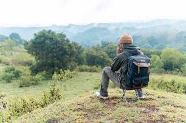 Bir adam kamp sandalyesine oturmuş, sırt çantası ve bir fincan kahveyle huzurlu dağ manzarasına hayran. Bir anlık yalnızlık, yansıma ve doğa anlayışı..