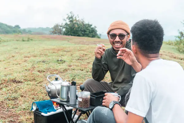 İki Endonezyalı adam kahve hazırlarken rahat bir ortamda birlikte gülüyorlar. Rahat bir kamp, kaynaşma ve çimenli bir arazide taşınabilir kahve takımlarıyla doğanın tadını çıkarma anı..
