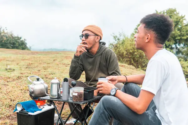 İki Endonezyalı adam kahve hazırlarken rahat bir ortamda birlikte gülüyorlar. Rahat bir kamp, kaynaşma ve çimenli bir arazide taşınabilir kahve takımlarıyla doğanın tadını çıkarma anı..