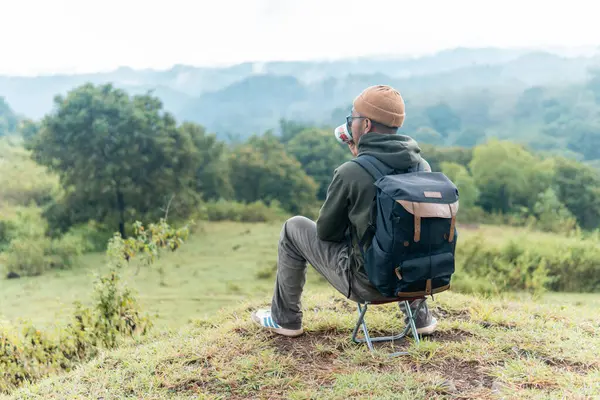 Bir adam kamp sandalyesine oturmuş, sırt çantası ve bir fincan kahveyle huzurlu dağ manzarasına hayran. Bir anlık yalnızlık, yansıma ve doğa anlayışı..