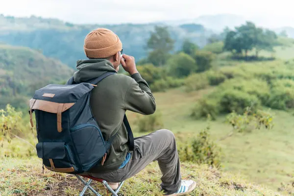 Bir adam kamp sandalyesine oturmuş, sırt çantası ve bir fincan kahveyle huzurlu dağ manzarasına hayran. Bir anlık yalnızlık, yansıma ve doğa anlayışı..