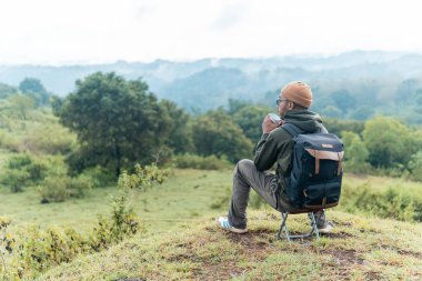 Bir adam kamp sandalyesine oturmuş, sırt çantası ve bir fincan kahveyle huzurlu dağ manzarasına hayran. Bir anlık yalnızlık, yansıma ve doğa anlayışı..