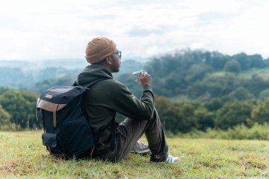 Endonezyalı bir adam sırt çantasıyla çimenlerde oturur, sakin bir dağ manzarasının tadını çıkarırken kahvesini yudumlar. Huzurlu yolculuk anları, yalnızlık ve doğa ile bağlantı yakalar..