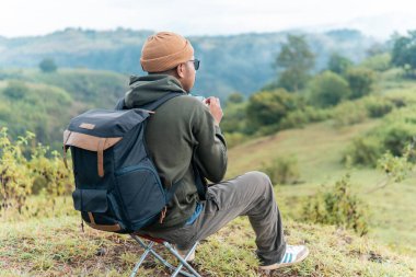 Bir adam kamp sandalyesine oturmuş, sırt çantası ve bir fincan kahveyle huzurlu dağ manzarasına hayran. Bir anlık yalnızlık, yansıma ve doğa anlayışı..