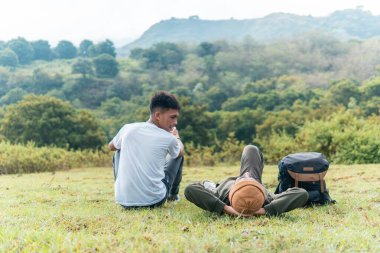 İki Endonezyalı adam çimenli bir tepede gülüyor ve manzaralı bir orman ve dağ manzaralı bir fincan kahvenin tadını çıkarıyor. Arkadaşlığı, özgürlüğü ve neşeli açık hava anlarını yakalar..