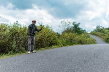 Sırt çantalı bir adam kırsal bir yolda otostop çekiyor. Yeşillik ve bulutlu gökyüzü ile çevrelenmiş bu görüntü tek başına seyahat etmenin, özgürlüğün ve vahşi doğada maceranın ruhunu yakalıyor..