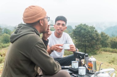 İki Endonezyalı adam kahve hazırlarken rahat bir ortamda birlikte gülüyorlar. Rahat bir kamp, kaynaşma ve çimenli bir arazide taşınabilir kahve takımlarıyla doğanın tadını çıkarma anı..