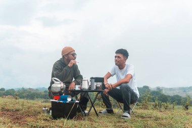 İki Endonezyalı adam kahve hazırlarken rahat bir ortamda birlikte gülüyorlar. Rahat bir kamp, kaynaşma ve çimenli bir arazide taşınabilir kahve takımlarıyla doğanın tadını çıkarma anı..