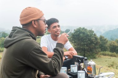 İki Endonezyalı adam kahve hazırlarken rahat bir ortamda birlikte gülüyorlar. Rahat bir kamp, kaynaşma ve çimenli bir arazide taşınabilir kahve takımlarıyla doğanın tadını çıkarma anı..
