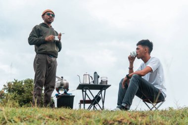 İki Endonezyalı adam kahve hazırlarken rahat bir ortamda birlikte gülüyorlar. Rahat bir kamp, kaynaşma ve çimenli bir arazide taşınabilir kahve takımlarıyla doğanın tadını çıkarma anı..