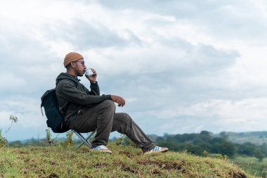 Bir adam kamp sandalyesine oturmuş, sırt çantası ve bir fincan kahveyle huzurlu dağ manzarasına hayran. Bir anlık yalnızlık, yansıma ve doğa anlayışı..