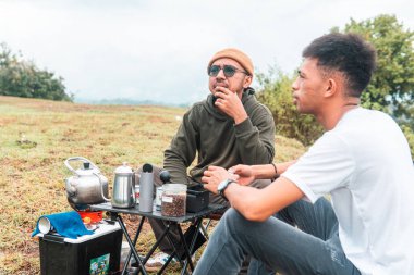 İki Endonezyalı adam kahve hazırlarken rahat bir ortamda birlikte gülüyorlar. Rahat bir kamp, kaynaşma ve çimenli bir arazide taşınabilir kahve takımlarıyla doğanın tadını çıkarma anı..