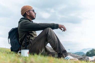 Endonezyalı bir adam sırt çantasıyla çimenlerde oturur, sakin bir dağ manzarasının tadını çıkarırken kahvesini yudumlar. Huzurlu yolculuk anları, yalnızlık ve doğa ile bağlantı yakalar..